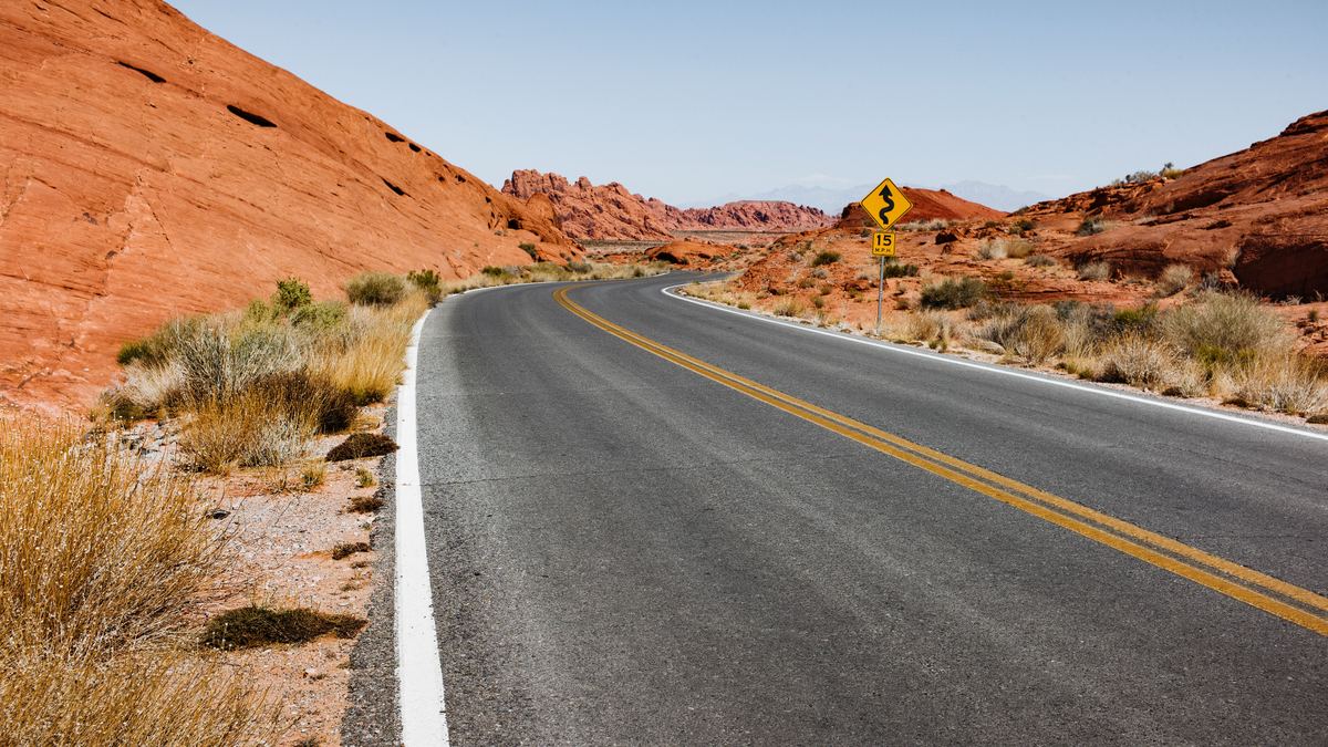 A paved road with a yellow center line curves through red sandstone hills and dry desert scrub under a clear blue sky.